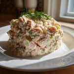 A mound of creamy salmon salad mixed with celery and topped with fresh dill, served on a white plate.
