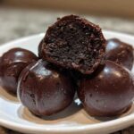 A stack of rich, dark chocolate covered oreo balls, with one cut in half showing the dense, truffle-like interior.