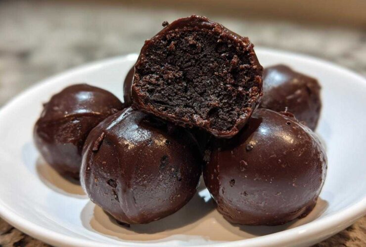 A stack of rich, dark chocolate covered oreo balls, with one cut in half showing the dense, truffle-like interior.