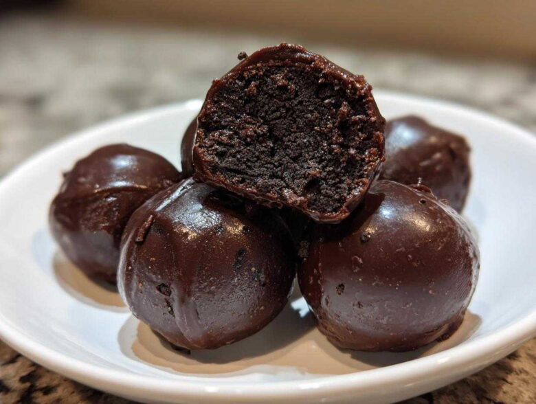 A stack of rich, dark chocolate covered oreo balls, with one cut in half showing the dense, truffle-like interior.