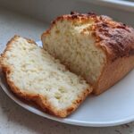 A loaf of golden-crusted beer bread, partially sliced, showing its soft, white interior on a white plate.