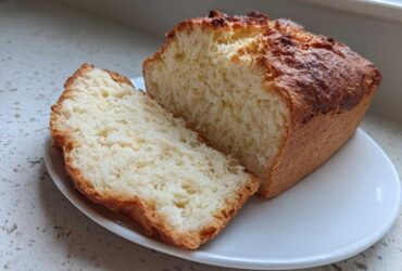 A loaf of golden-crusted beer bread, partially sliced, showing its soft, white interior on a white plate.