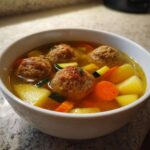 A close-up of a white bowl filled with flavorful albondigas soup, featuring meatballs, potatoes, carrots, and zucchini.