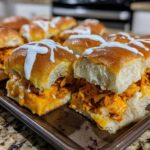 A pan full of freshly baked buffalo chicken sliders topped with melted cheese and a white ranch or blue cheese drizzle.