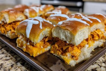 A pan full of freshly baked buffalo chicken sliders topped with melted cheese and a white ranch or blue cheese drizzle.
