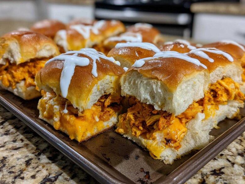A pan full of freshly baked buffalo chicken sliders topped with melted cheese and a white ranch or blue cheese drizzle.