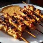 Close-up of five grilled chicken satay skewers on a white plate next to a dollop of peanut dipping sauce.
