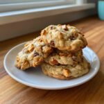A stack of four soft, chewy kitchen sink cookies loaded with chocolate chips and caramel pieces on a white plate.