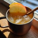 A close-up of an apple cider moscow mule served over ice in a copper mug, garnished with apple slices and a cinnamon stick.