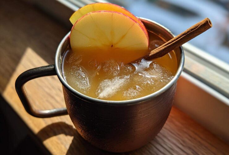 A close-up of an apple cider moscow mule served over ice in a copper mug, garnished with apple slices and a cinnamon stick.