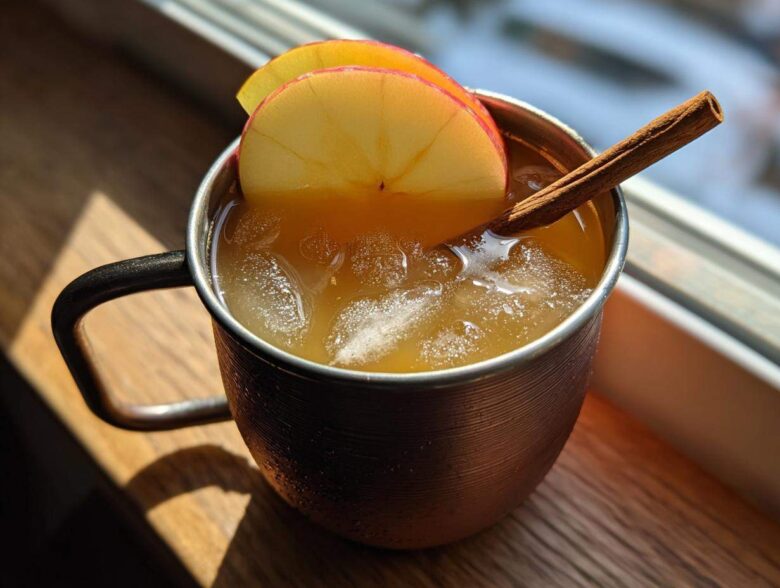 A close-up of an apple cider moscow mule served over ice in a copper mug, garnished with apple slices and a cinnamon stick.