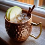 Close-up of a chilled apple cider moscow mule in a hammered copper mug, garnished with green apple slices and cinnamon sticks.