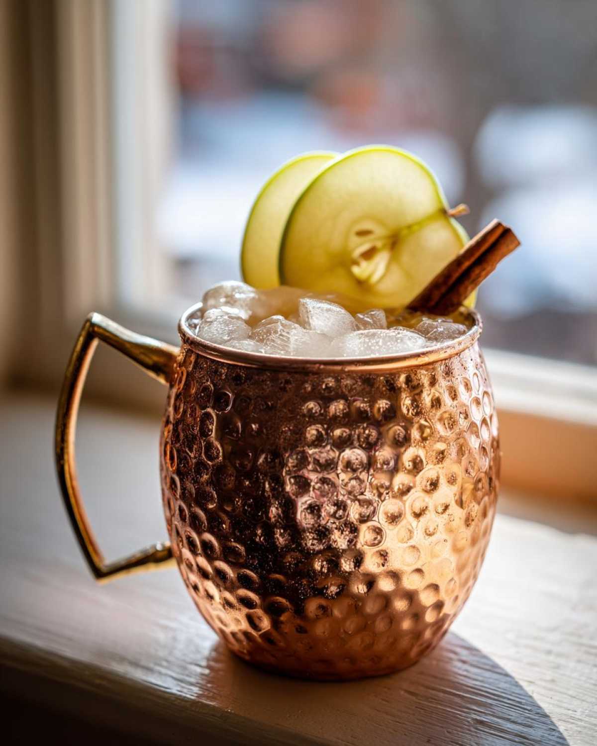 A refreshing apple cider moscow mule served over ice in a hammered copper mug, garnished with apple slices and a cinnamon stick.