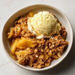 A close-up of warm apple crisp topped with a scoop of melting vanilla ice cream in a light-colored bowl.