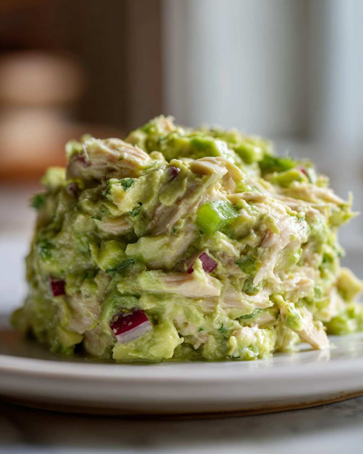 A mound of creamy avocado chicken salad mixed with shredded chicken, red onion, and green onion pieces on a white plate.