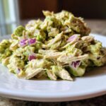 A close-up serving of creamy avocado chicken salad mixed with shredded chicken and diced red onion on a white plate.