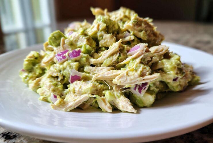 A close-up serving of creamy avocado chicken salad mixed with shredded chicken and diced red onion on a white plate.