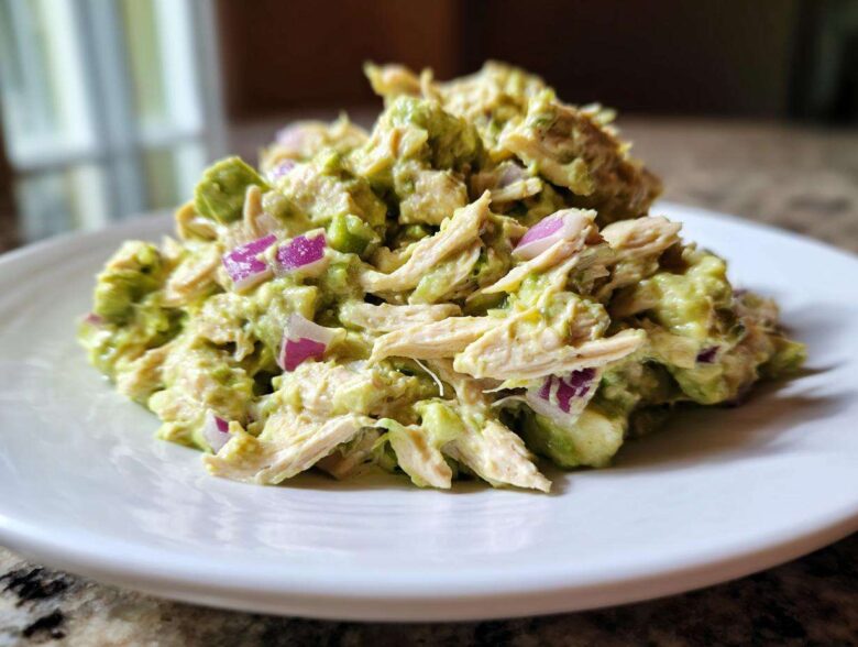 A close-up serving of creamy avocado chicken salad mixed with shredded chicken and diced red onion on a white plate.