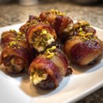 Close-up of several bacon wrapped stuffed dates, filled with cheese and topped with chopped pistachios.