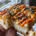 A hand holding up a thick, square portion of baked sushi bake topped with bright orange spicy mayo and seaweed flakes.