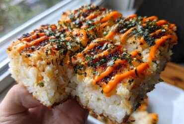A hand holding up a thick, square portion of baked sushi bake topped with bright orange spicy mayo and seaweed flakes.