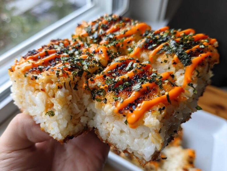 A hand holding up a thick, square portion of baked sushi bake topped with bright orange spicy mayo and seaweed flakes.