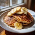 A stack of golden brown banana oatmeal pancakes topped with fresh banana slices and syrup.