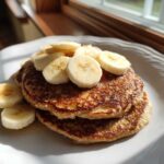 A stack of golden brown banana oatmeal pancakes topped with fresh banana slices, drizzled with syrup.