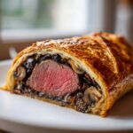 Close-up of a slice of beef wellington recipe showing medium-rare tenderloin wrapped in golden puff pastry and duxelles.