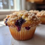Close-up of a golden blueberry muffin featuring a thick, sugary crumb topping and bursting blueberries.