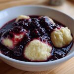 Close-up of fluffy dumplings served in a white bowl covered with rich, dark blackberry sauce.