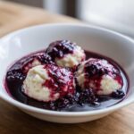 Close-up of three fluffy Blackberry Dumplings swimming in a deep purple blackberry sauce in a white bowl.