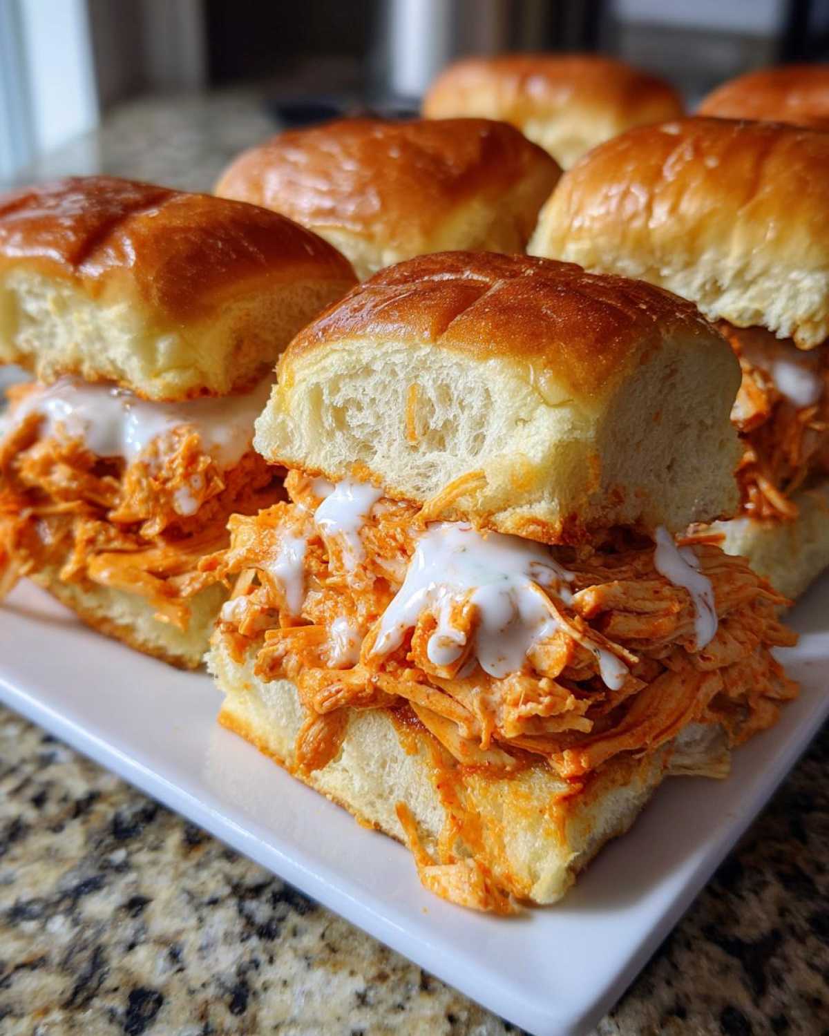 Close-up of several buffalo chicken sliders piled high with shredded orange chicken and drizzled with ranch dressing.