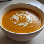 A close-up of a bowl of vibrant orange butternut squash soup, garnished with a swirl of cream.
