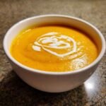 A close-up of vibrant orange butternut squash soup topped with a swirl of cream in a white bowl.