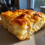 A close-up, golden-brown slice of cheesy potato casserole with a bubbly, melted cheese topping.