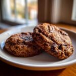 Close-up of two rich, dark vegan chocolate chip cookies resting on a light plate near a window.