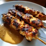 Three skewers of perfectly grilled, slightly charred chicken satay served next to a pool of peanut dipping sauce.