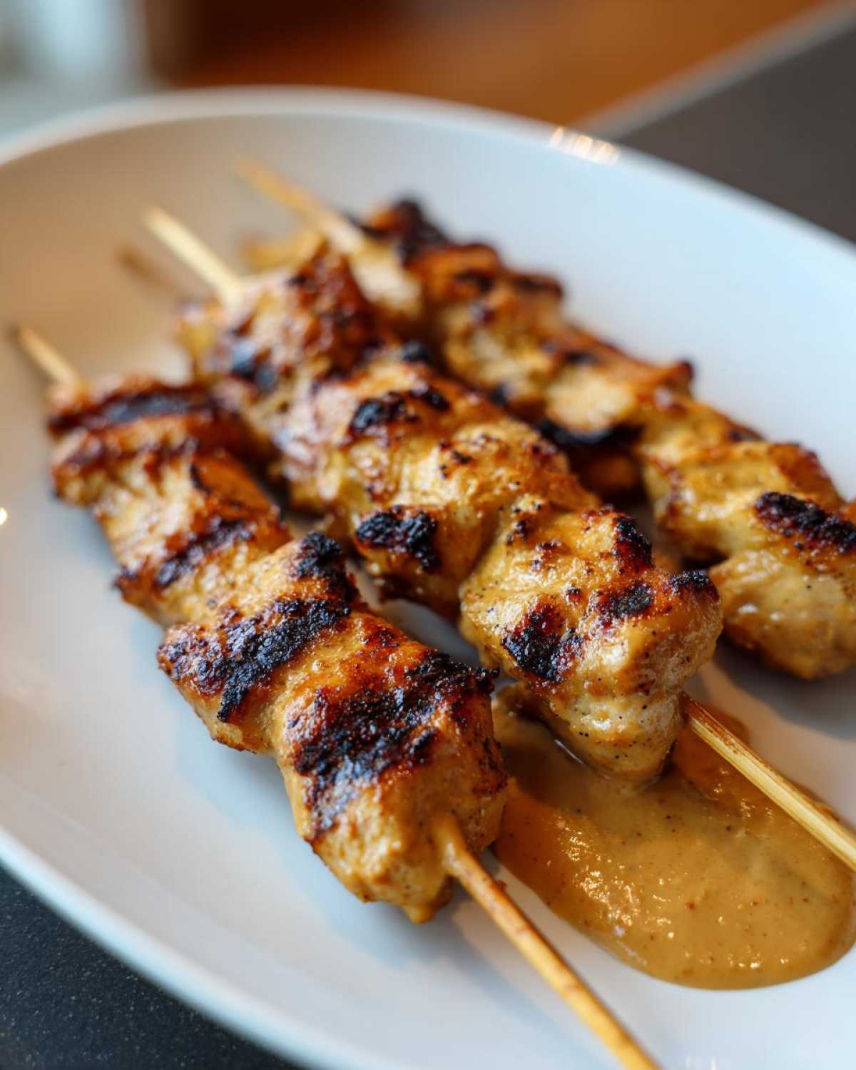 Three skewers of grilled chicken satay with visible char marks resting on a white plate with peanut sauce.