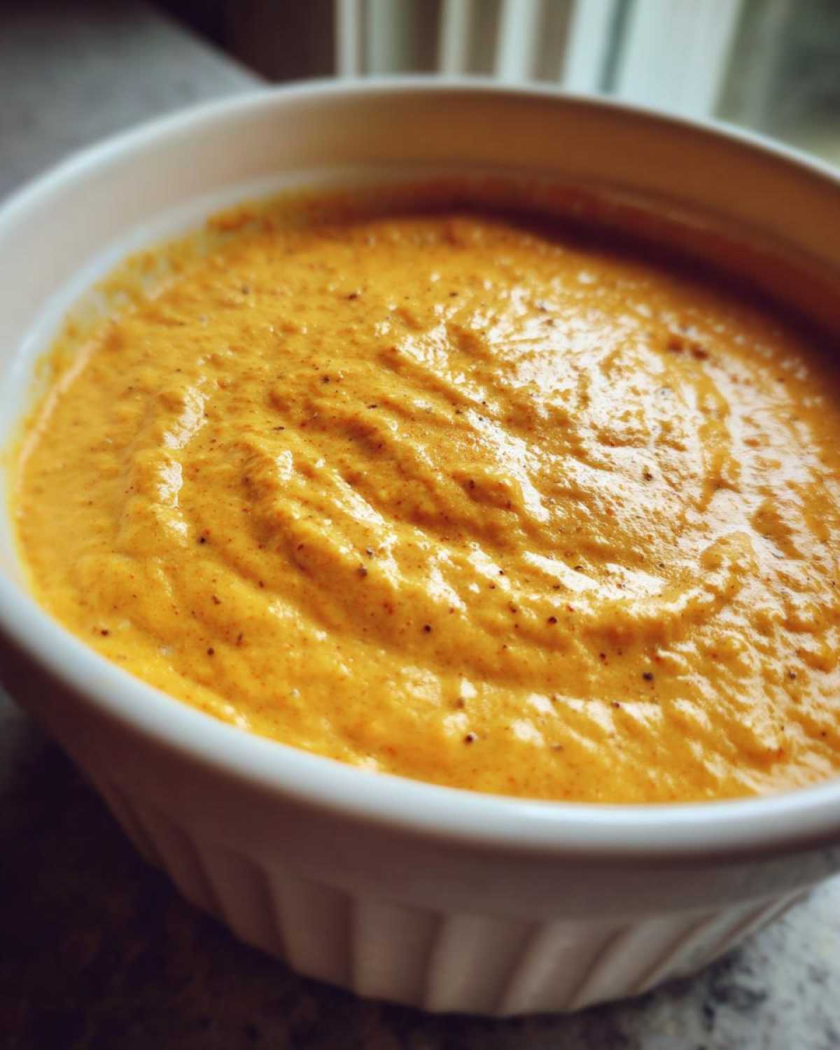 Close-up of thick, orange-yellow beer cheese dip with visible spices in a white fluted ramekin.