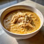 A close-up of a bowl of creamy chicken chili recipe, topped with shredded white cheese and pulled chicken.