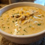 Close-up of a creamy chicken chili recipe served hot in a white bowl, topped with shredded cheese and parsley.