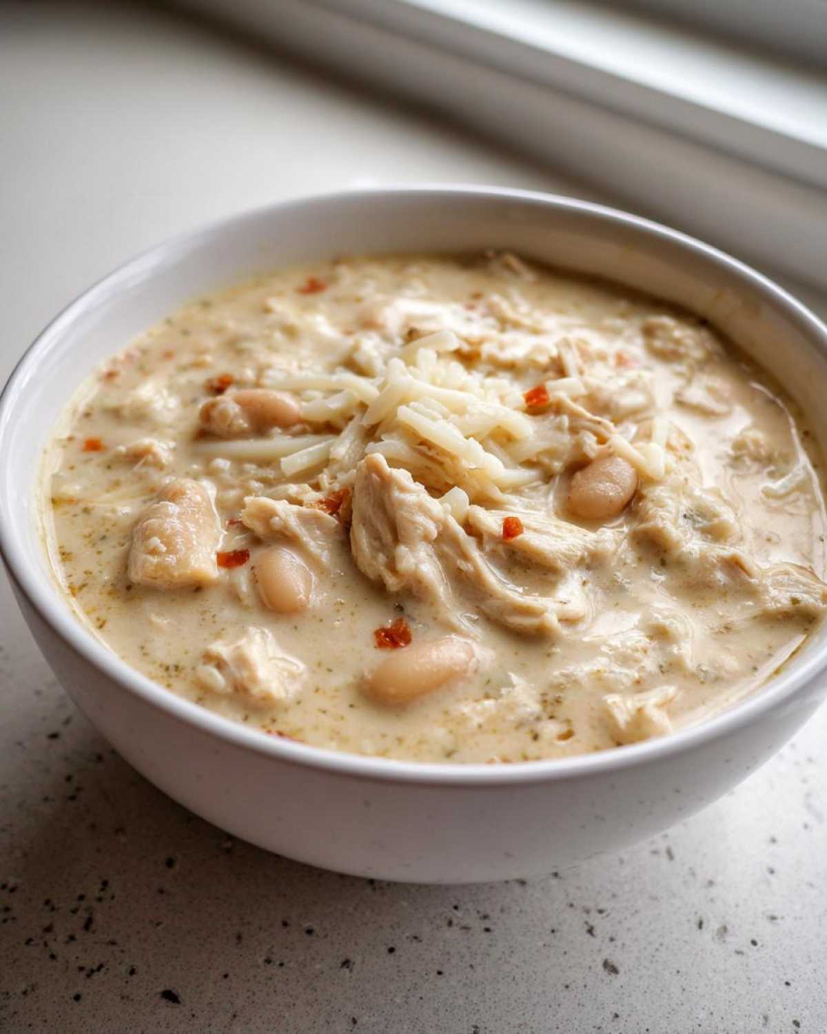 Close-up of a white bowl filled with creamy chicken chili recipe, topped with shredded cheese.