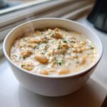 Close-up of a steaming white bowl filled with creamy chicken chili, featuring shredded chicken, white beans, and green herbs.