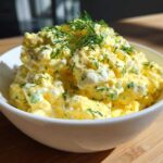 A white bowl filled high with creamy, yellow egg salad recipe, topped with fresh dill.