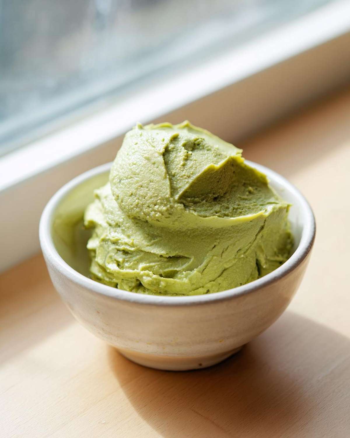 A scoop of vibrant green, creamy olive dip served in a rustic white bowl near a window.