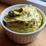 A close-up of thick, vibrant green olive dip served in a small white fluted ramekin on a wooden surface.