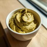 Close-up of a thick, creamy green olive dip swirled high in a small white bowl, brightly lit by sunlight.