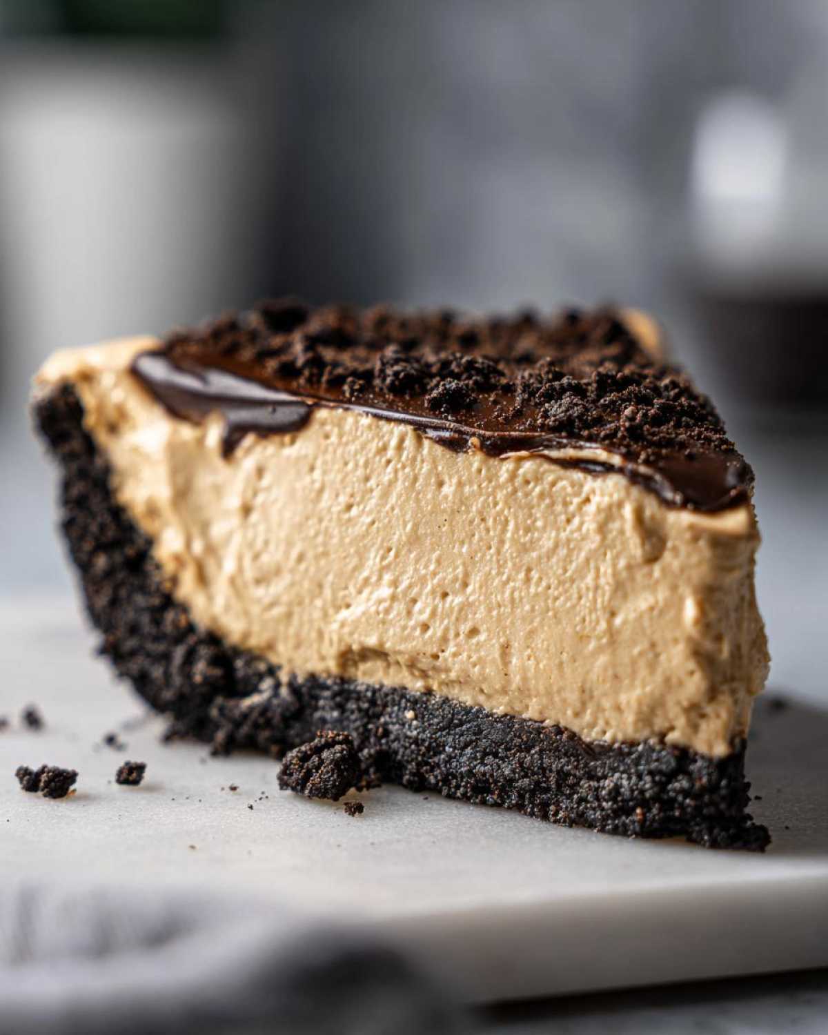 A close-up slice of creamy peanut butter pie with a dark chocolate cookie crust, topped with chocolate ganache and crumbles.