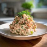 A mound of creamy salmon salad mixed with celery and topped with fresh dill, served on a white plate.